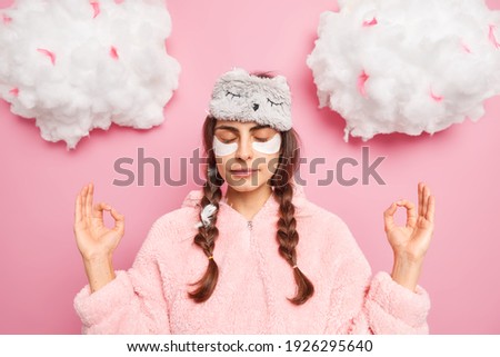 Relaxed beautiful woman in pajamas meditates after awakening keeps hands in zen gesture closes eyes enjoys peaceful atmosphere undergoes beauty treatments at home practices yoga stands indoor