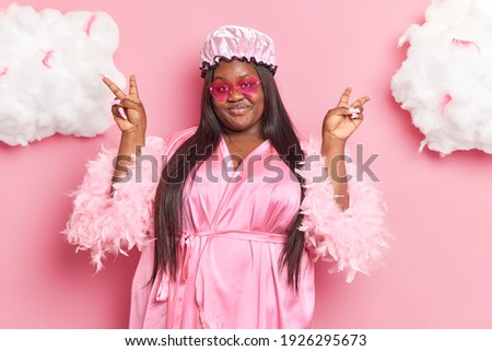 Smiling glad overweight woman with dark skin makes peace gesture with both hands has long nails dressed in casual domestic clothes makes victory sign isolated over pink background clouds above