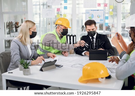 Competent architects, designers and engineers in medical masks planning and creating construction project during working meeting. Business during pandemic. Cooperation between diverse people.