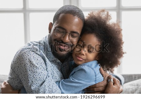 Portrait of happy cute Black girl hugging her beloved daddy and smiling with closed eyes. Peaceful affectionate dad and daughter kid enjoying time together, embracing, relaxing at home. Close up
