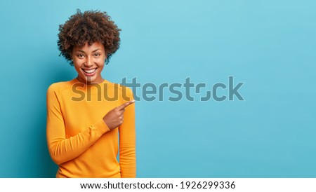 Smiling black curly haired woman in casual jumper points aside shows space for your advertisement or promo promots big sale has special offer isolated over blue background. Great suggestion.