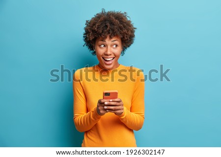 Positive curly haired ethnic woman uses mobile phone checks messages and reads news holds modern cellular in hands looks with curious happy expression on right isolated over blue background. Royalty-Free Stock Photo #1926302147