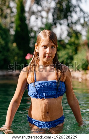 A beautiful little girl with long dark hair in a blue swimsuit swims in the outdoor pool on a hot sunny day. Summer vacation. Selective focus, defocus.