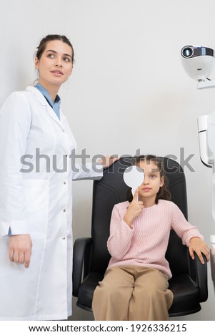 Cute little girl in casualwear doing eye test in modern ophthalmological clinics while young female optician in whitecoat standing near by