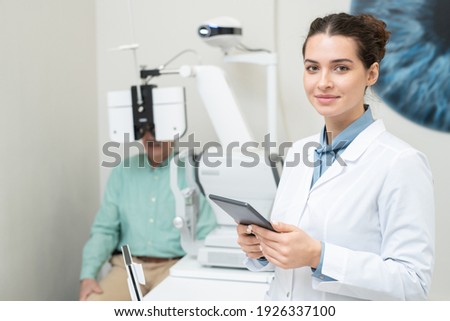 Happy young female ophthalmologist in whitecoat looking at you while using tablet on background of patient having his eyesight checked up