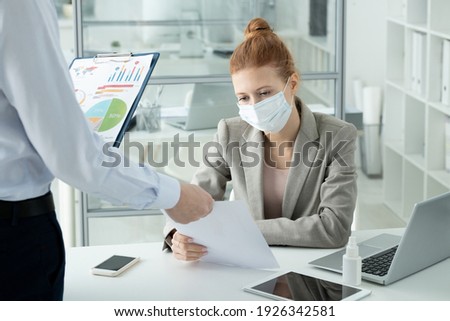 Young serious female office worker in protective mask and formalwear looking through paper held by her colleague while sitting by desk