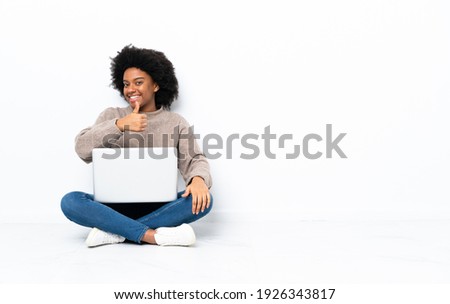 Young African American woman with a laptop sitting on the floor giving a thumbs up gesture