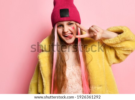 Close up portrait of an active and cheerful teenage girl who is showing V-sign with her fingers and tongue. Little rebel in a pink hat, yellow fur coat posing on a pink background. Adolescence concept