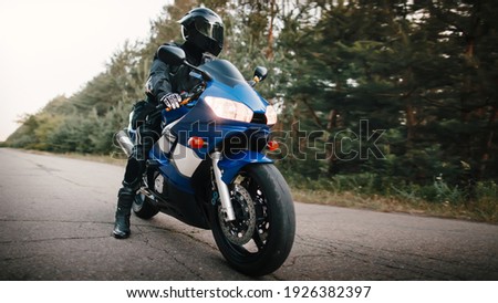 Motorcyclist in leather protective suit and black helmet sits on sports motorcycle. Biker in black rides on the road against the background of the forest Royalty-Free Stock Photo #1926382397