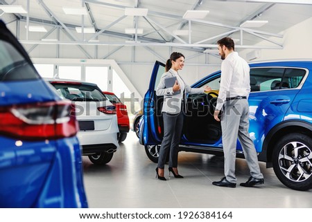 Friendly, smiling female seller showing brand new car to a customer while standing in car salon. Royalty-Free Stock Photo #1926384164