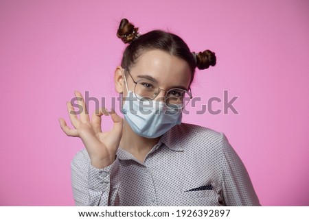 Modern responsible young girl in protective breathing antibacterial medical facial mask showing okay gesture looking at camera, preventing world coronavirus out spread on pink studio background.