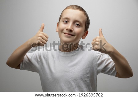 Happy candid child boy showing thumbs up, like hands gesture. Positive satisfied kid recommending advertising service or product looking at camera on gray studio background with copy space for ad.