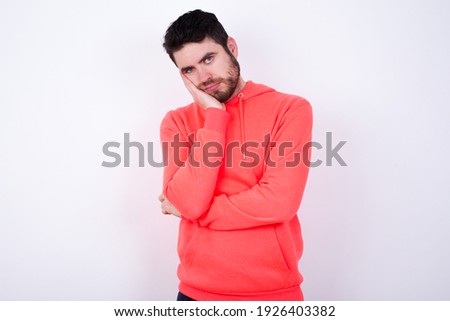 Very bored young Caucasian bearded man wearing pink hoodie against white background holding hand on cheek while support it with another crossed hand, looking tired and sick.