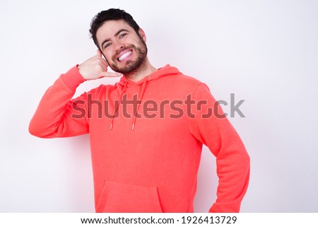 young Caucasian bearded man wearing pink hoodie against white background imitates telephone conversation, makes phone call gesture with hands, has confident expression. Call me!