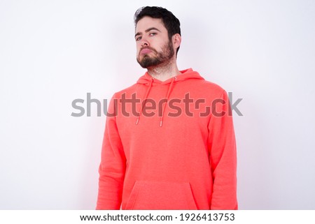 young Caucasian bearded man wearing pink hoodie against white background with snobbish expression curving lips and raising eyebrows, looking with doubtful and skeptical expression, suspect and doubt. Royalty-Free Stock Photo #1926413753