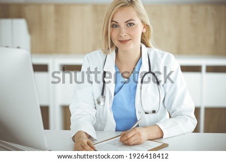 Portrate of woman-doctor at work while sitting at the desk in clinic. Blonde cheerful physician filling up medical form or prescription