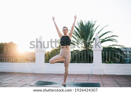 Full length of young slender barefooted female standing in Tree pose with raised arms gesturing mudra sign during yoga session on villa rooftop at sunset