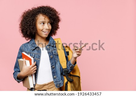 Smiling little clever african kid school girl 12-13 year old in casual clothes backpack hold books point index finger aside on copy space isolated on pastel pink background Childhood education concept