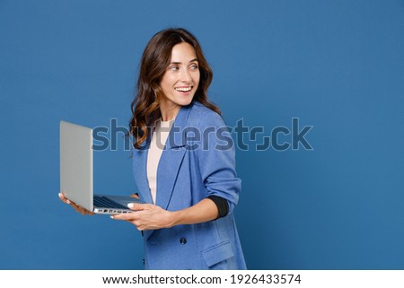 Smiling cheerful beautiful attractive young brunette woman 20s wearing basic jacket standing working on laptop pc computer looking aside isolated on bright blue colour wall background studio portrait Royalty-Free Stock Photo #1926433574
