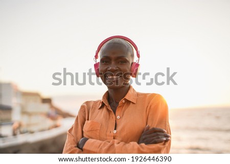 Happy senior african woman looking in camera while wearing headphones and listening music form playlist - Beach and sunset in background