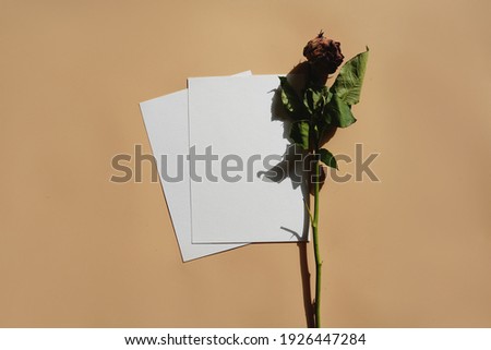 An empty sheet of white paper and a dried rose on a pastel yellow surface. A blank greeting card, mock up.