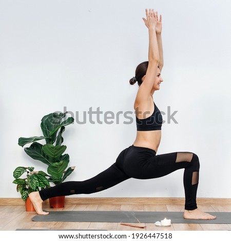 Smiling young woman practicing yoga holding warrior asana with arms outstretched up. In a bright room. Burning incense stick aroma and candle. She's happy and smiling