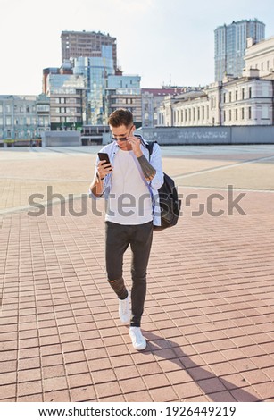 A young guy, European appearance, in sunglasses and with a black backpack. Standing on a city street and looking into the phone.