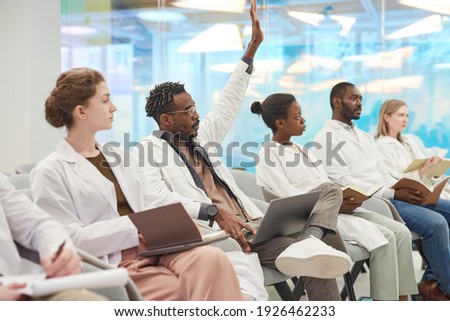 Multi-ethnic group of people wearing lab coats sitting in row in audience at medical seminar, focus on African American man raising hand to ask question, copy space Royalty-Free Stock Photo #1926462233