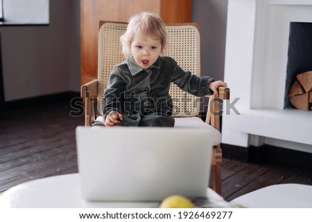 Cute little blonde girl watching cartoons on her laptop. The child smiles in a cheerful mood. He plays his own games. Children's clothing green dress and boots.