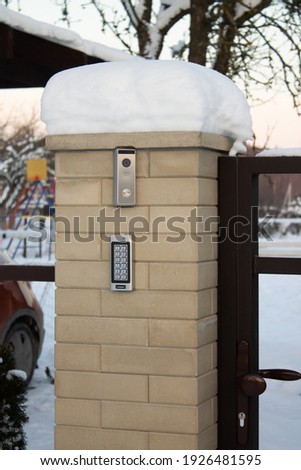 Silver intercom call panel with blue number buttons and a video camera, on a brick beige fence pillar of a private house. Vertical
