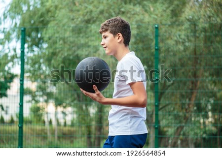  Cute Teenager in white t-shirt with orange basketball ball plays basketball on street playground in summer. Hobby, active lifestyle, sports activity for kids.	
