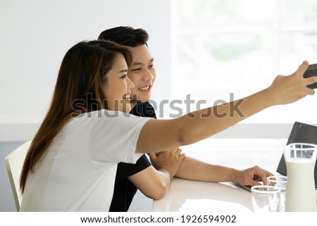 Young asian couple sit together taking selfie using mobile phone. Glasses of milk on desk. Stay home togetherness concept.