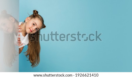 Happy kid, girl on blue studio background. Looks happy, cheerful. Copyspace for ad. Childhood, education, emotions, business, facial expression concept. Peeking out from behind the wall. Flyer
