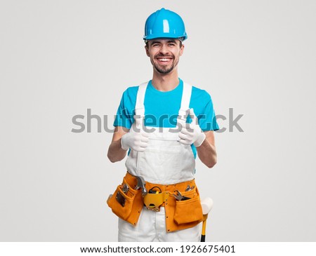 Positive handyman in overall and hardhat with toolkit showing thumbs up and smiling brightly against white background