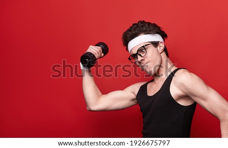 Side view of comic skinny young male in sportswear and eyeglasses making funny serious face while doing bicep exercise with dumbbell on red background