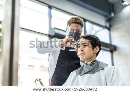 Professional male stylist cutting man's hair in salon. The man wearing mask and face shield to prevent from coronavirus infection during pandemic. New normal beauty salon or barber business concept. Royalty-Free Stock Photo #1926680492