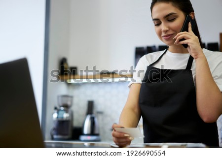 Professional manager of restaurant enjoying job making mobile phone call to delivery service, positive 20s woman barista making booking via cellular holding visit card standing at working space
