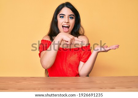 Brunette teenager girl wearing casual clothes sitting on the table amazed and smiling to the camera while presenting with hand and pointing with finger. 