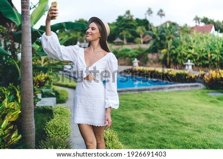 Positive female in white dress smiling and taking picture on mobile phone while standing in tropical garden on resort on summer day
