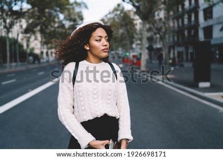 Calm African American female in casual clothes and beret with backpack walking on street after hard workday and looking away