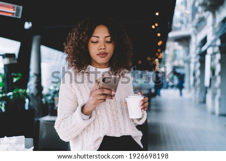 Young African American female in casual outfit standing at street cafe and browsing mobile phone with takeaway coffee in hand