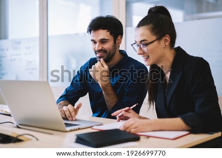 Cheerful young multiethnic corporate lawyers in formal wear editing text of new contract using laptop sitting at table with notebook in modern office