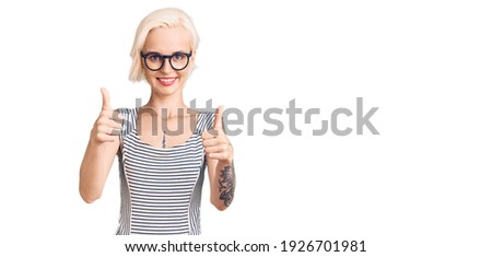 Young blonde woman with tattoo wearing casual clothes and glasses success sign doing positive gesture with hand, thumbs up smiling and happy. cheerful expression and winner gesture. 