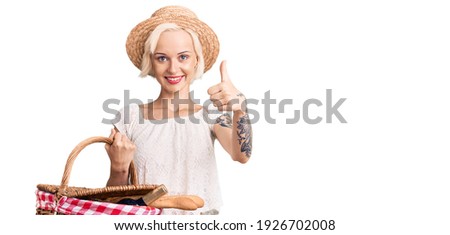 Young blonde woman with tattoo wearing summer hat and holding picnic wicker basket with bread smiling happy and positive, thumb up doing excellent and approval sign 