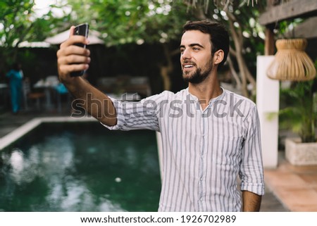 Happy young bearded male blogger in stylish clothes taking selfie on mobile phone while relaxing at poolside in contemporary hotel