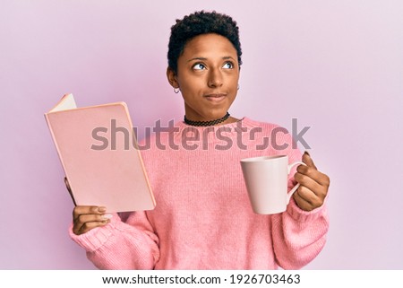 Young african american girl reading a book and drinking a cup of coffee smiling looking to the side and staring away thinking. 