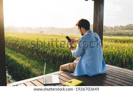 Back view of male freelancer in casual outfit using smartphone while working on laptop remotely in wooden gazebo surrounded green area