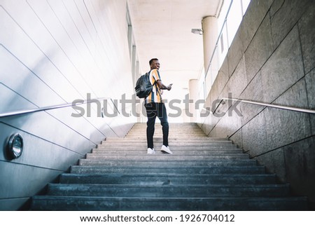 Cheerful male tourist dressed in stylish fashion outfit smiling during sightseeing time in travel city, back view of happy African American student with trendy backpack climb urban stairs Royalty-Free Stock Photo #1926704012