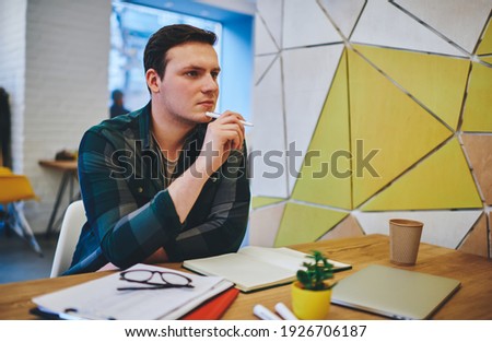 Serious male in casual clothes looking away sitting at table with notebook thinking about idea for new project working in light workspace