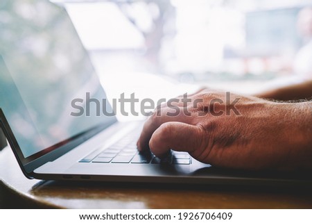 Side view of crop anonymous male sitting at table near window touching keyboard of laptop while working in modern workspace